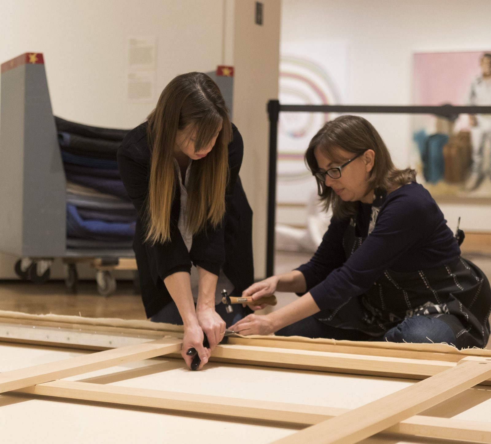 Two women attach a canvas to a large frame